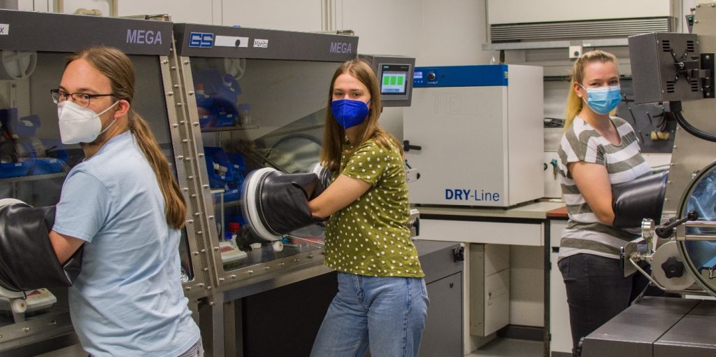 Three people in a lab with their hands in glove-boxes, all wearing masks.