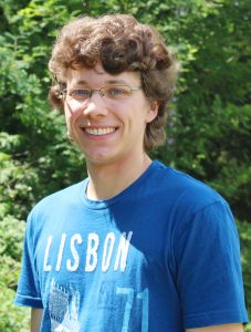 Jakob smiling in the camera. He has curly brown hair and has glasses on.